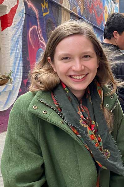 A young woman standing outside in front of a wall, which is covered in graffiti