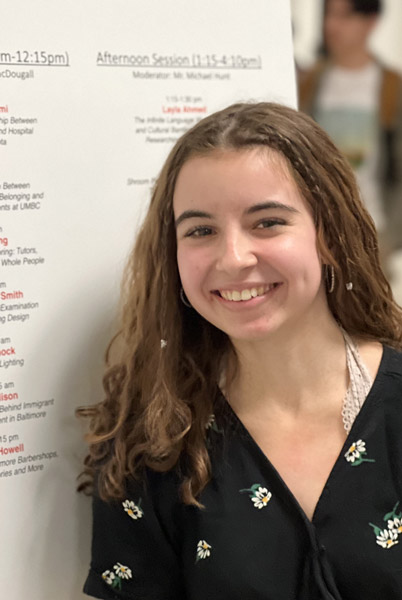 A young female undergraduate student standing in front of a placard that features her work at a research presentation event on campus